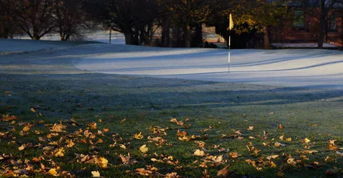 This landscape photograph shows a frozen golf course on a winter morning in Tapton Park, Chesterfield, United Kingdom. The ground is covered with a layer of frost, and autumn leaves are scattered across the grass in the foreground, highlighting the seasonal transition. The image captures the nature of the park with its mature trees providing a scenic background, while the golf flag stands out as the main feature on the frosty course. The low morning sunlight casts long shadows from the trees, enhancing the wintry atmosphere typical of this time of year.
